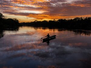 A fisherman rows a boat across the Amazon River at sunset, surrounded by vivid reflections.