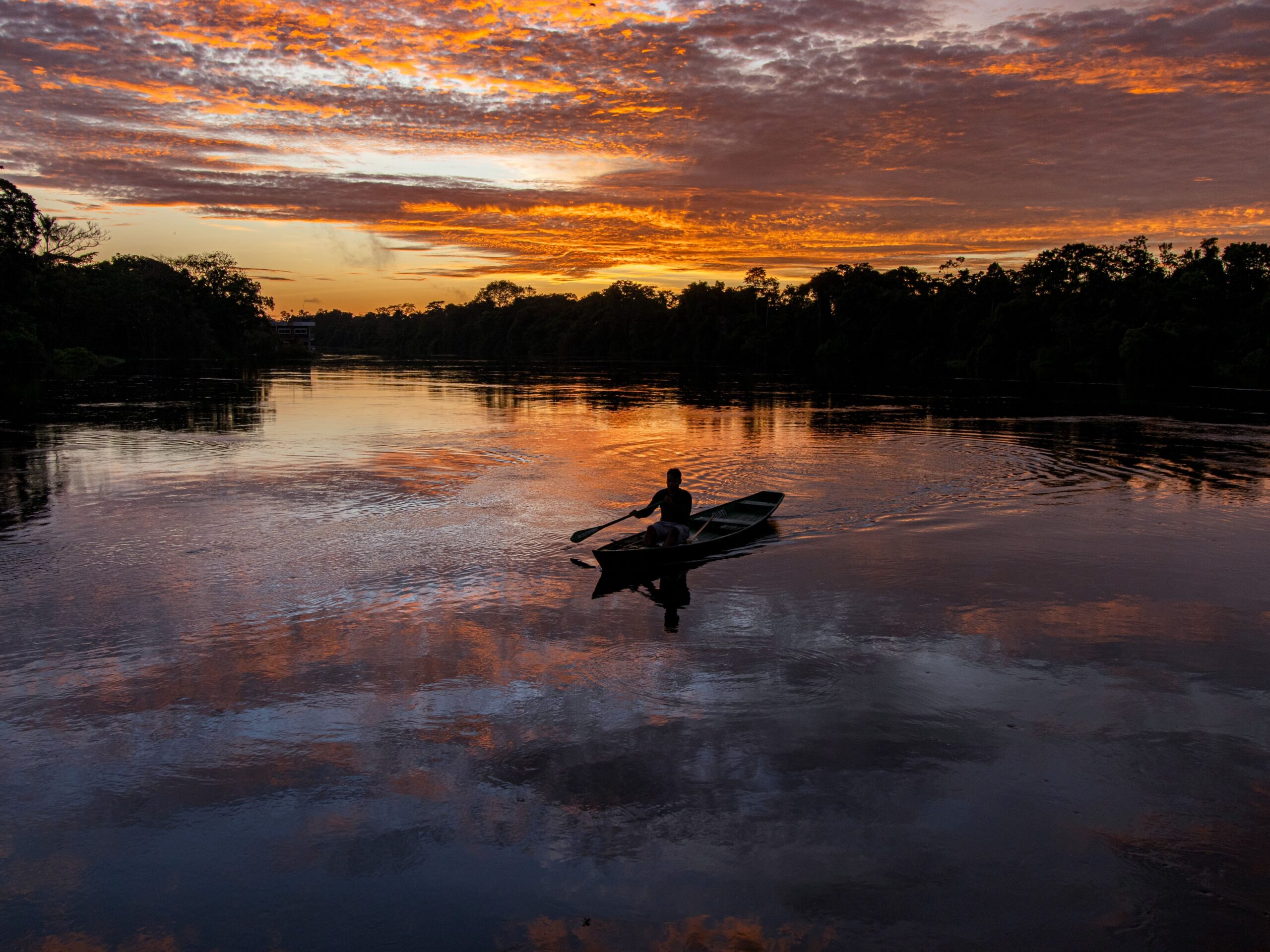 A fisherman rows a boat across the Amazon River at sunset, surrounded by vivid reflections.